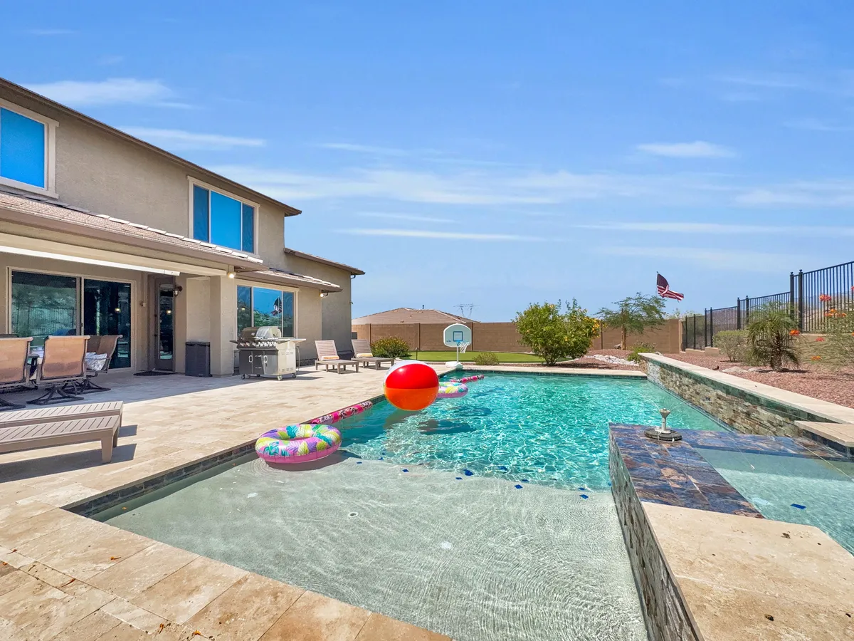 A cheerful backyard scene featuring a swimming pool, a colorful beach ball floating in the water, poolside lounge chairs, and a covered back patio. The lively ambiance suggests opportunities for poolside games, barbecues, and gathering with friends or family.