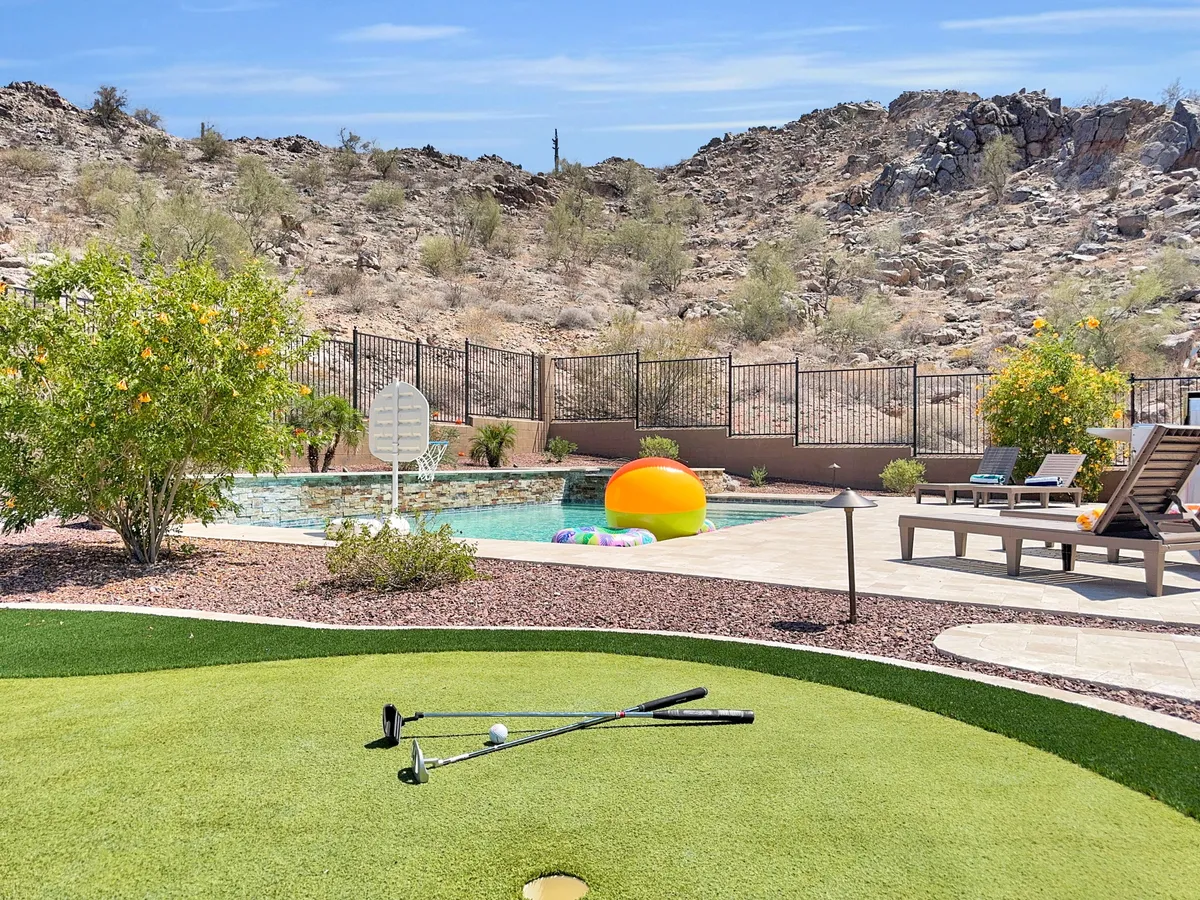 This playful exterior shot displays a golf putting green, swimming pool with floaties, green lawn, and landscaped plants, with golf clubs and ball in the foreground. The scene suggests leisurely putting, poolside fun, and outdoor games with a scenic mountain view.