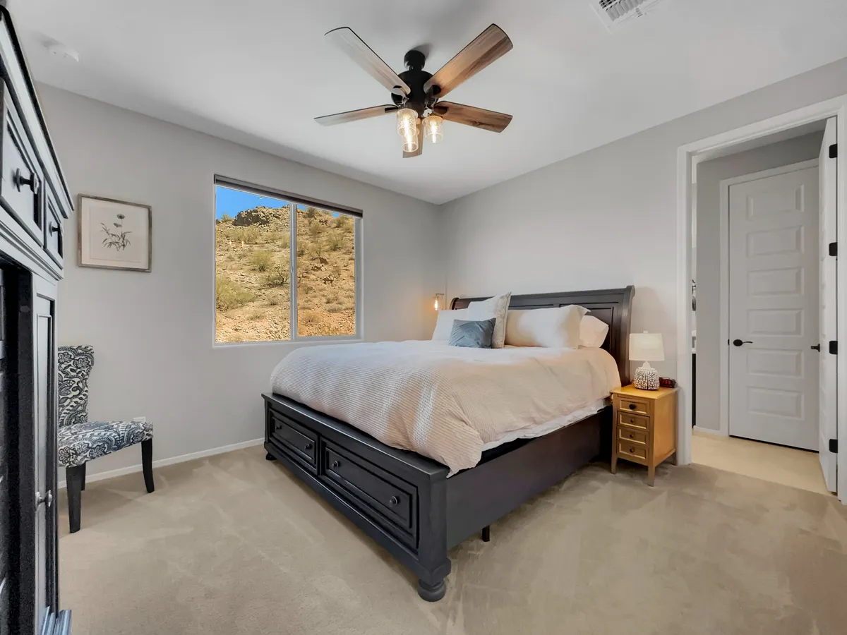 The queen bedroom features a cozy, queen-sized bed facing a large window with a stunning mountain view, complemented by a decorative chair and a bedside lamp for nighttime reading. The airy space and soft textiles invite relaxation and accessibility.
