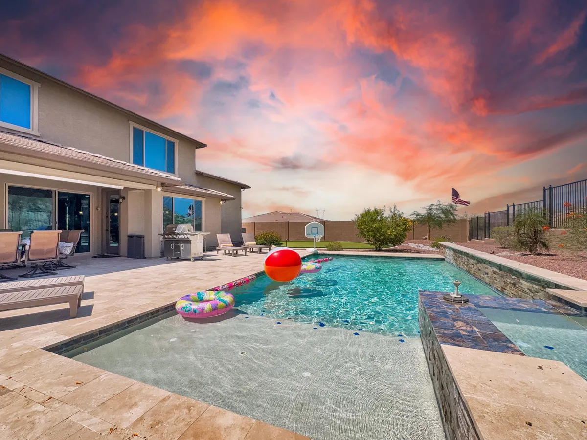 A vibrant backyard scene featuring a sparkling swimming pool with colorful pool floats, a back patio seating area, and a dramatic sunset sky over the landscape. The area suggests fun, relaxation, and outdoor enjoyment, surrounded by scenic views.
