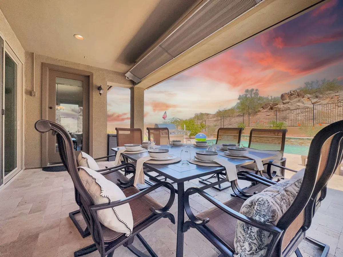 This exterior shot showcases an elegant outdoor dining table and a pool area set against a dramatic sunset sky and mountainous backdrop. The setup suggests al fresco dining and evening social gatherings as the sun sets over the desert landscape.