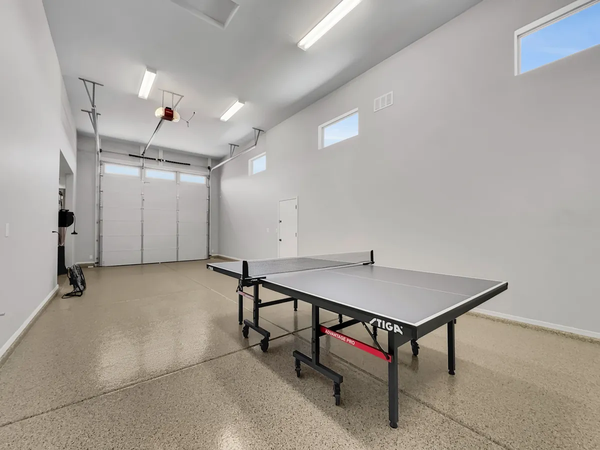 This image captures a ping pong table within a large garage, highlighted by a prominent garage door and bright walls, reinforcing the flexibility and scale of the space. The open setting suggests the garage can accommodate vehicles or serve as a gathering point for social events, aligning with the idea of ample parking and group activities.