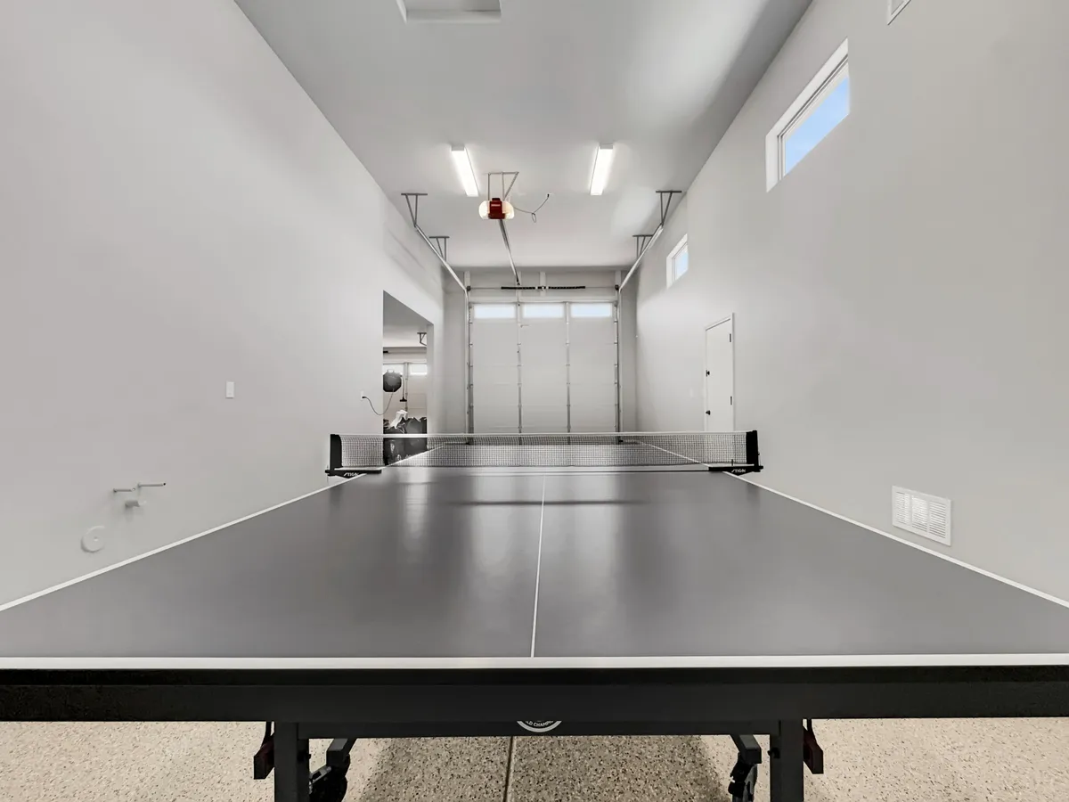 This image features a ping pong table set up in a spacious garage with bright overhead lighting and a visible garage door, suggesting a multi-functional space often associated with RV storage or large-scale parking. The open, energetic setup emphasizes ample room for recreational activities, as well as the functional aspect of garage space.
