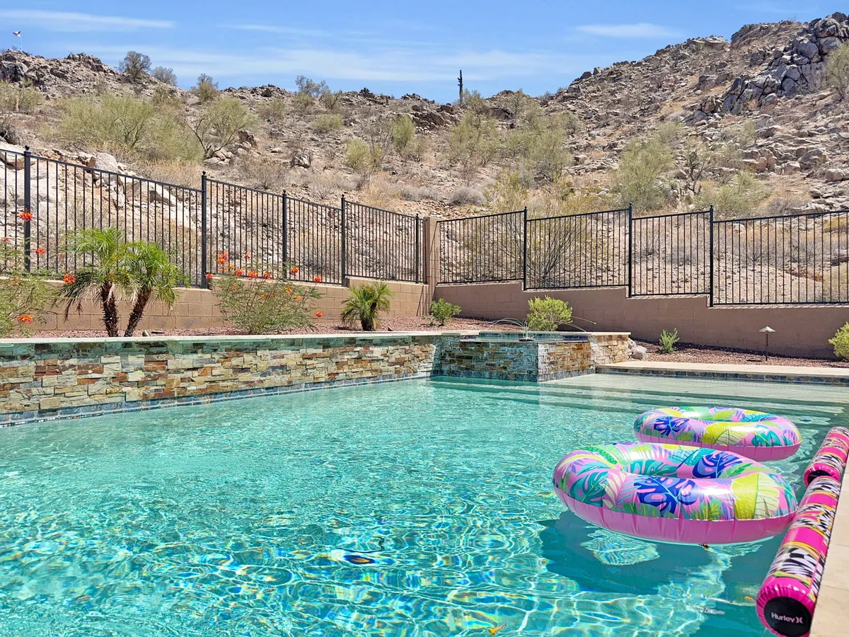 An inviting swimming pool accented by colorful pool floats, set against a dramatic rocky landscape and mountain views. A secure fence encloses the area, providing both safety and privacy while enhancing the tranquil atmosphere. The surroundings capture the beauty of the Arizona outdoors.