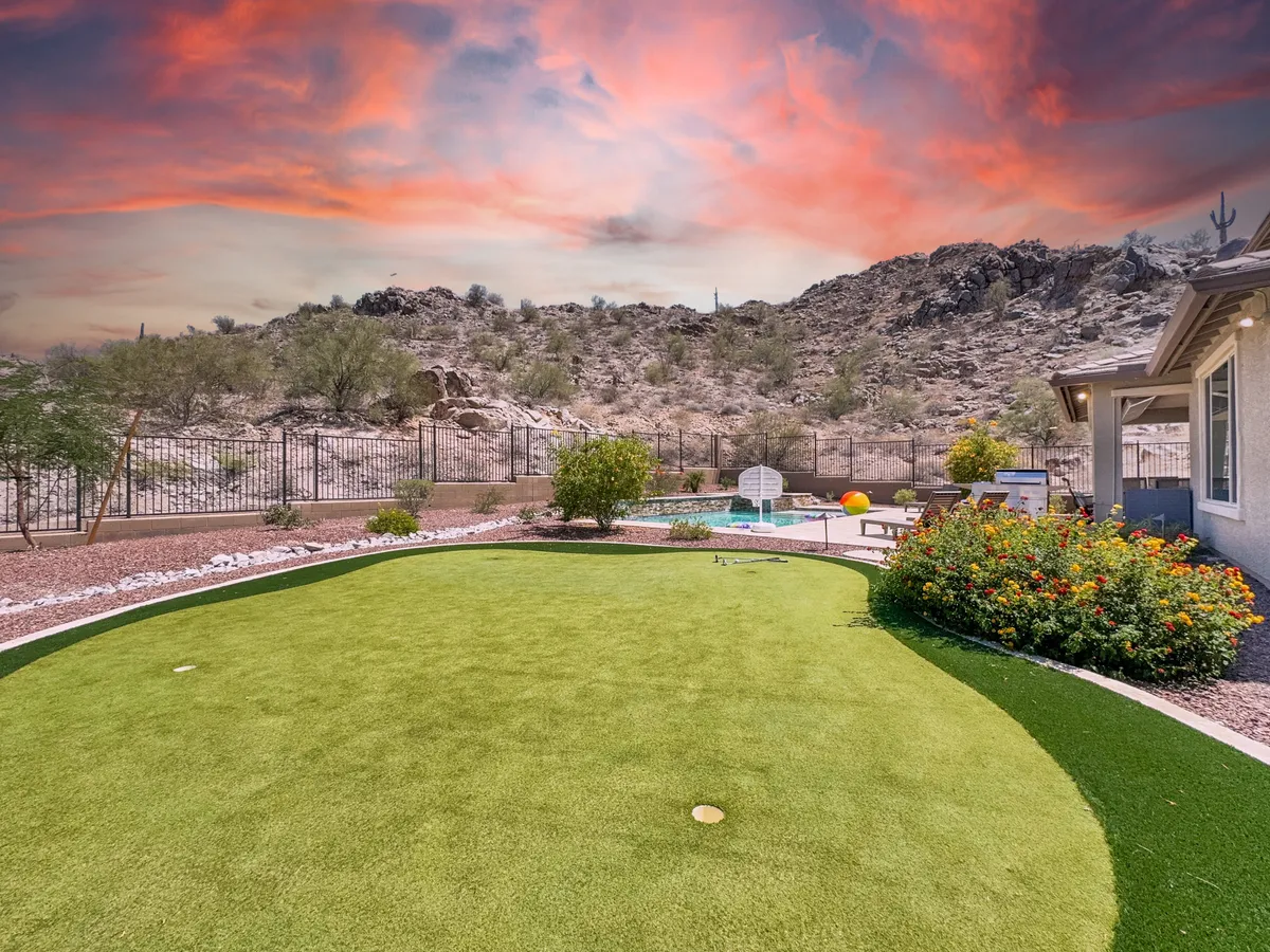 This exterior photo features a tranquil pool area with artificial grass, cozy seating, and a striking mountainous backdrop under a vibrant sky. The image highlights the natural beauty surrounding the home and the inviting outdoor setting.