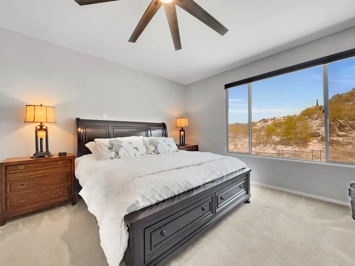 A beautifully appointed king-sized bed is the centerpiece of this tranquil bedroom, complete with decorative pillows, a large window offering natural light and a view, and inviting nightstands with softly glowing lamps. The room exudes a peaceful, hotel-like ambiance with its neutral tones and plush bedding.
